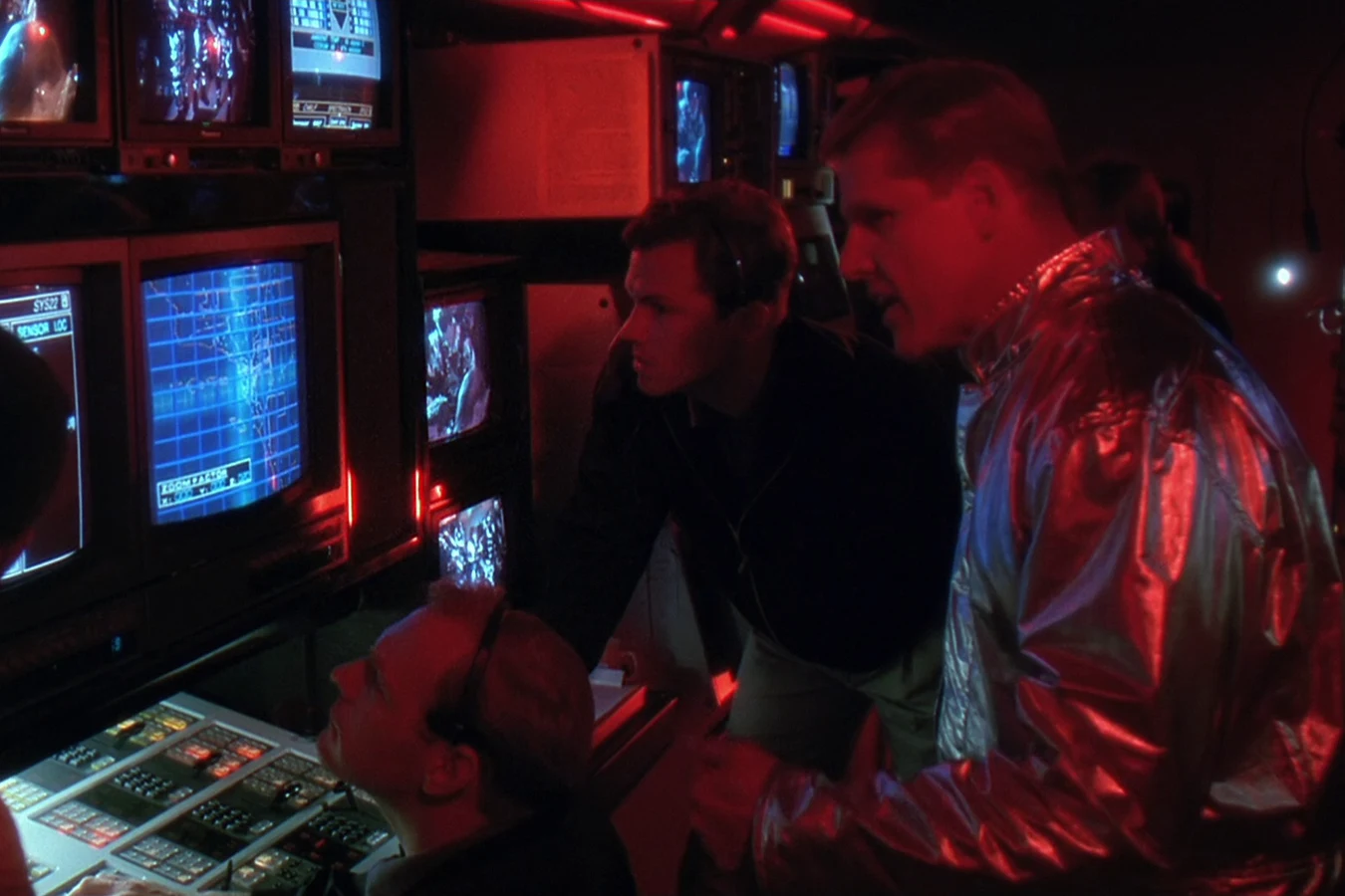 Garber wearing a headset beside Peter Keyes in a silver suit among the monitors of the red-lit OWLF control room in Predator 2