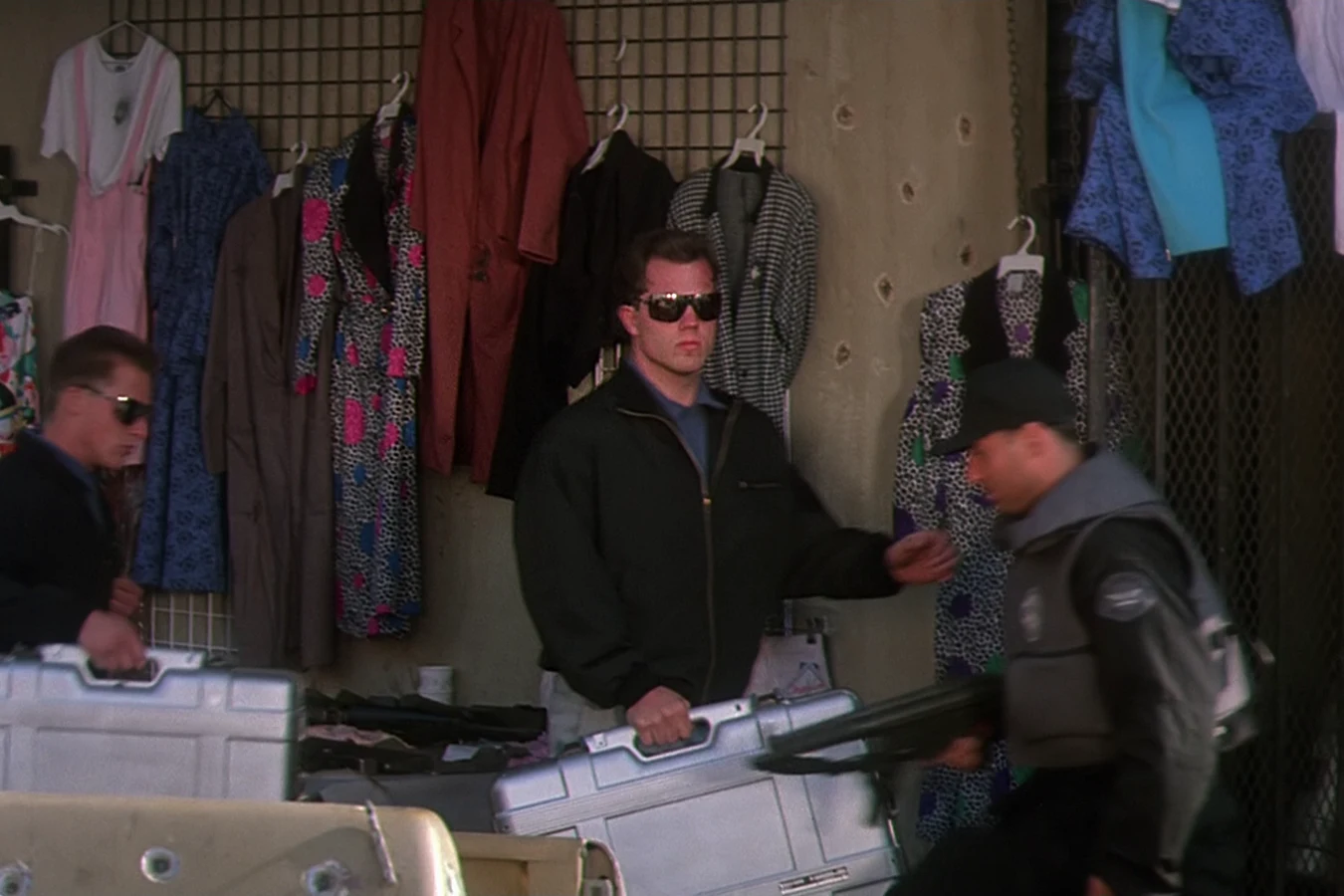 Garber in sunglasses and a dark jacket carrying a silver case past racks of clothes at a market stall in Predator 2