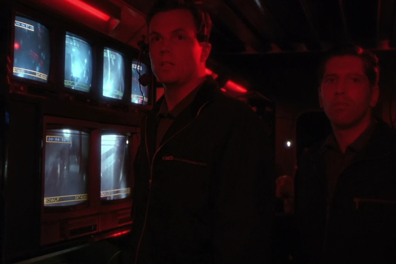 Garber in a headset standing beside another agent in front of OWLF surveillance monitors in red light in Predator 2