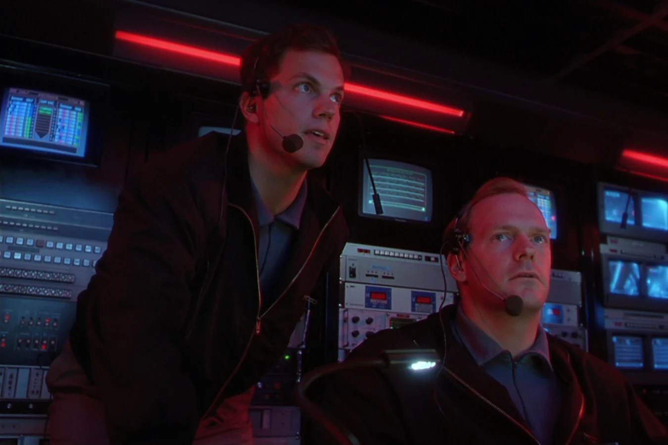 Garber wearing a headset and looking up at the monitors beside a seated agent in the OWLF control room in Predator 2