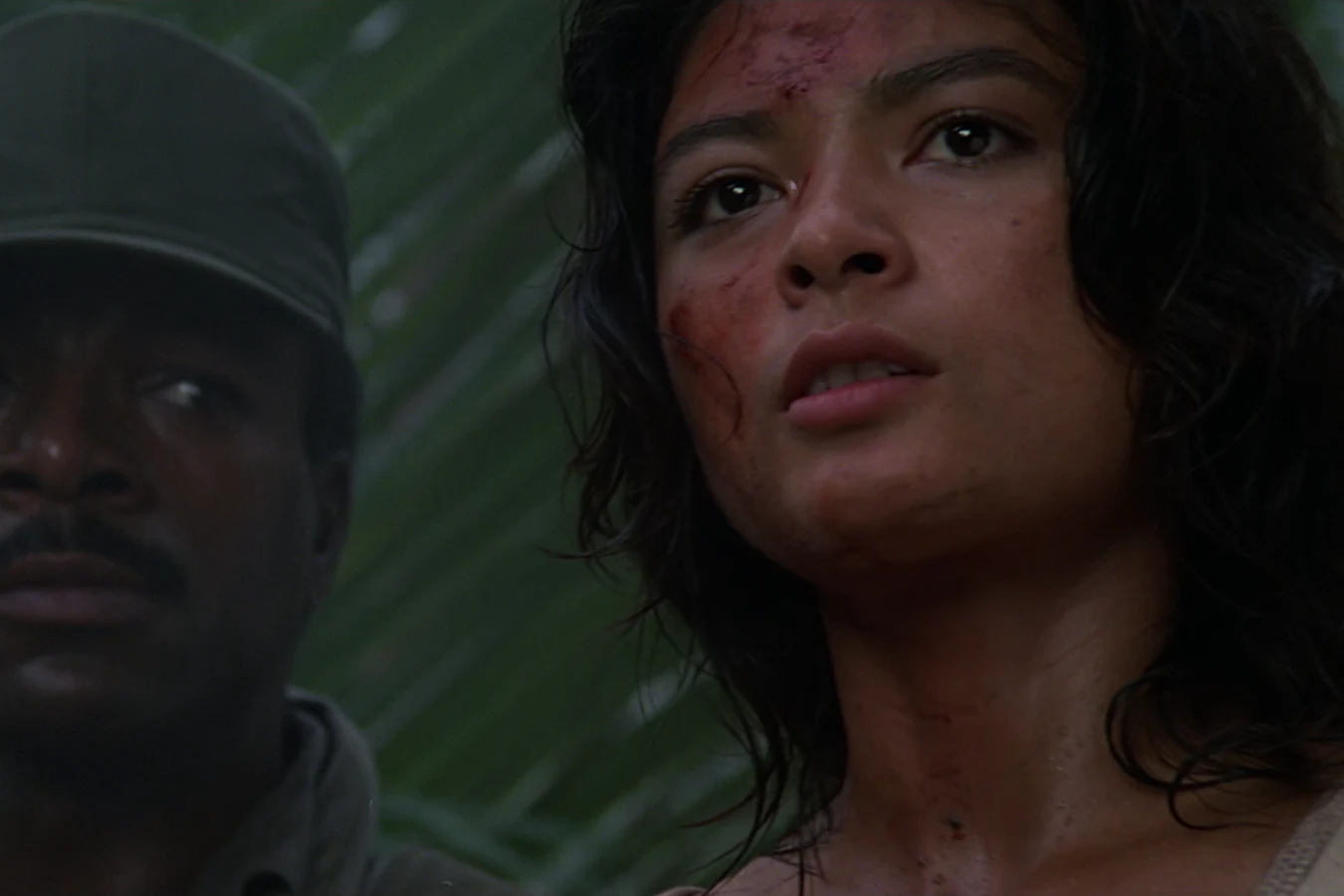 Close-up of Anna Gonsalves with a cut on her forehead, with Dillon watching beside her in the jungle