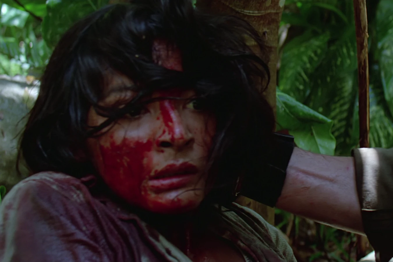 Anna Gonsalves crouching in the jungle with her face and shirt drenched in Hawkins' blood