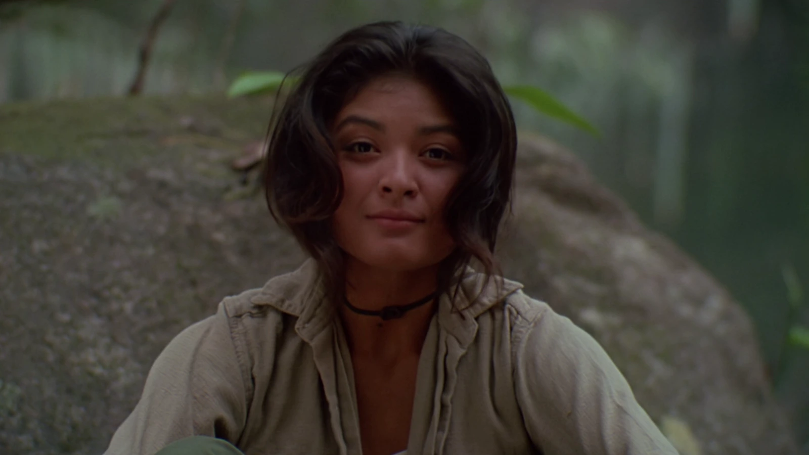 Anna Gonsalves with a faint smile, sitting in front of a large mossy rock in the jungle