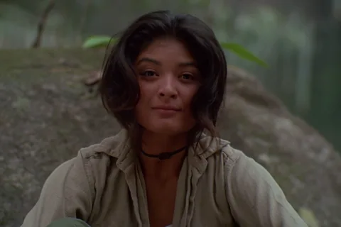 Anna Gonsalves with a faint smile, sitting in front of a large mossy rock in the jungle