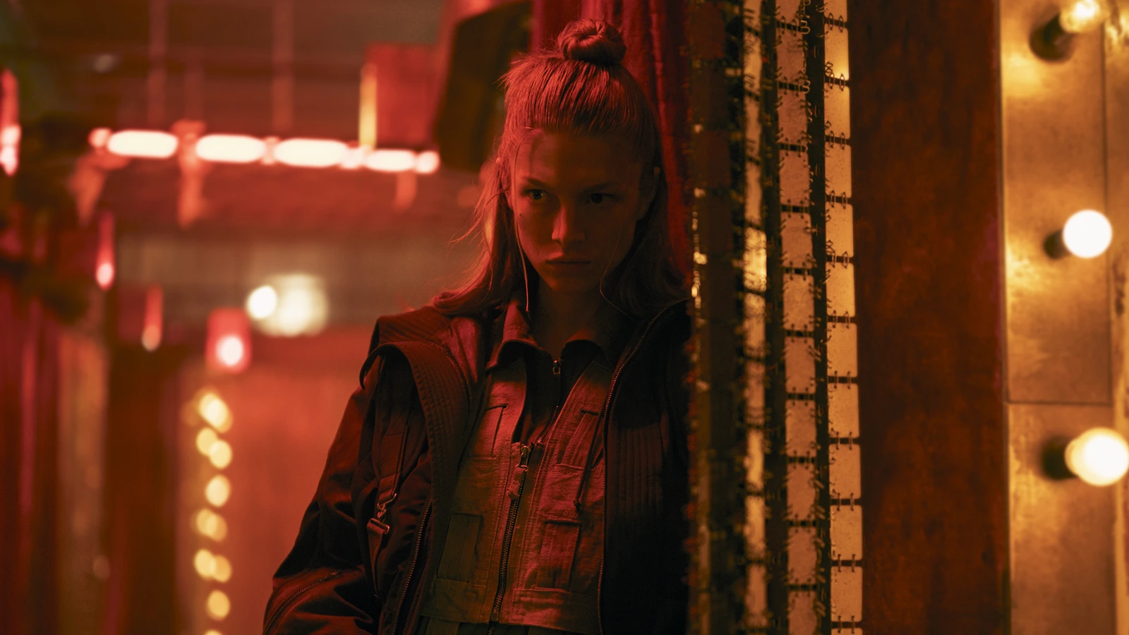Hunter Schafer as Cora in red light, standing by a bulb-lined pillar in Blade Runner 2099