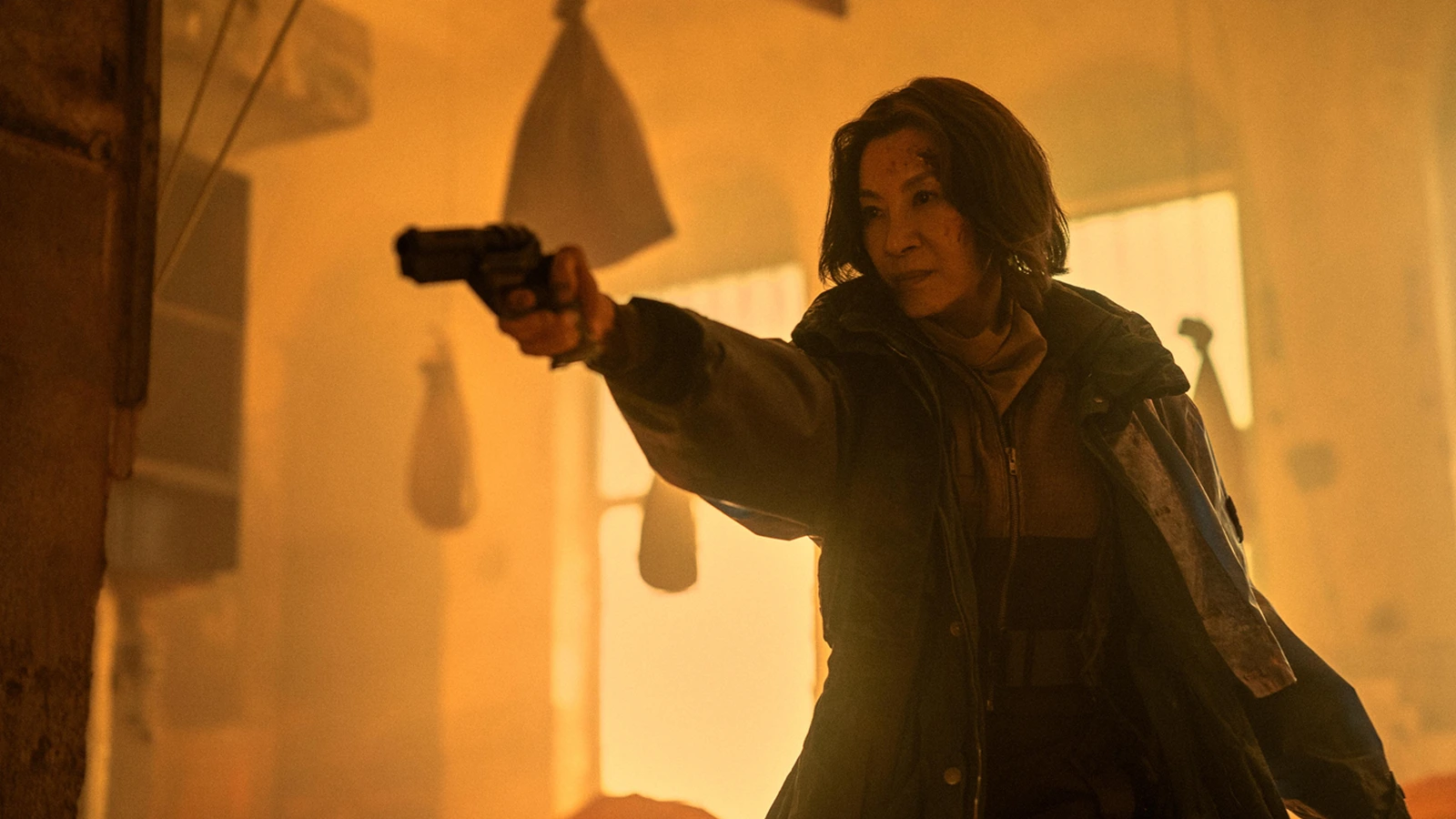 Michelle Yeoh as Olwen aiming a pistol in amber light, with a scratch on her cheek, in Blade Runner 2099