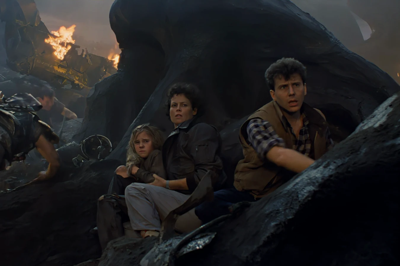 Carter Burke, Ellen Ripley, and Newt sheltering in the rocks beside the burning dropship at Hadley's Hope in Aliens