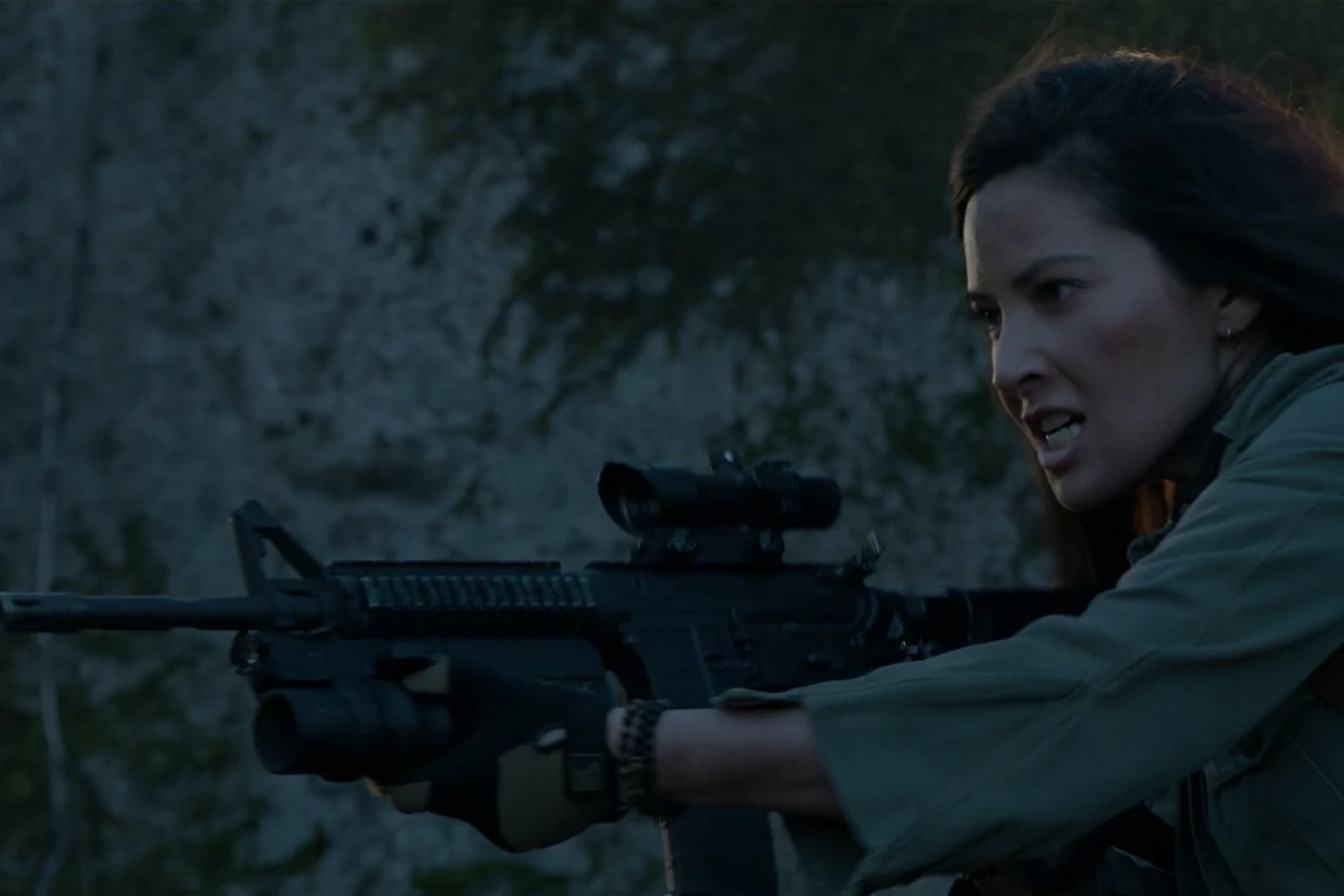 Casey Brackett aiming a scoped rifle in the woods in front of a rock wall