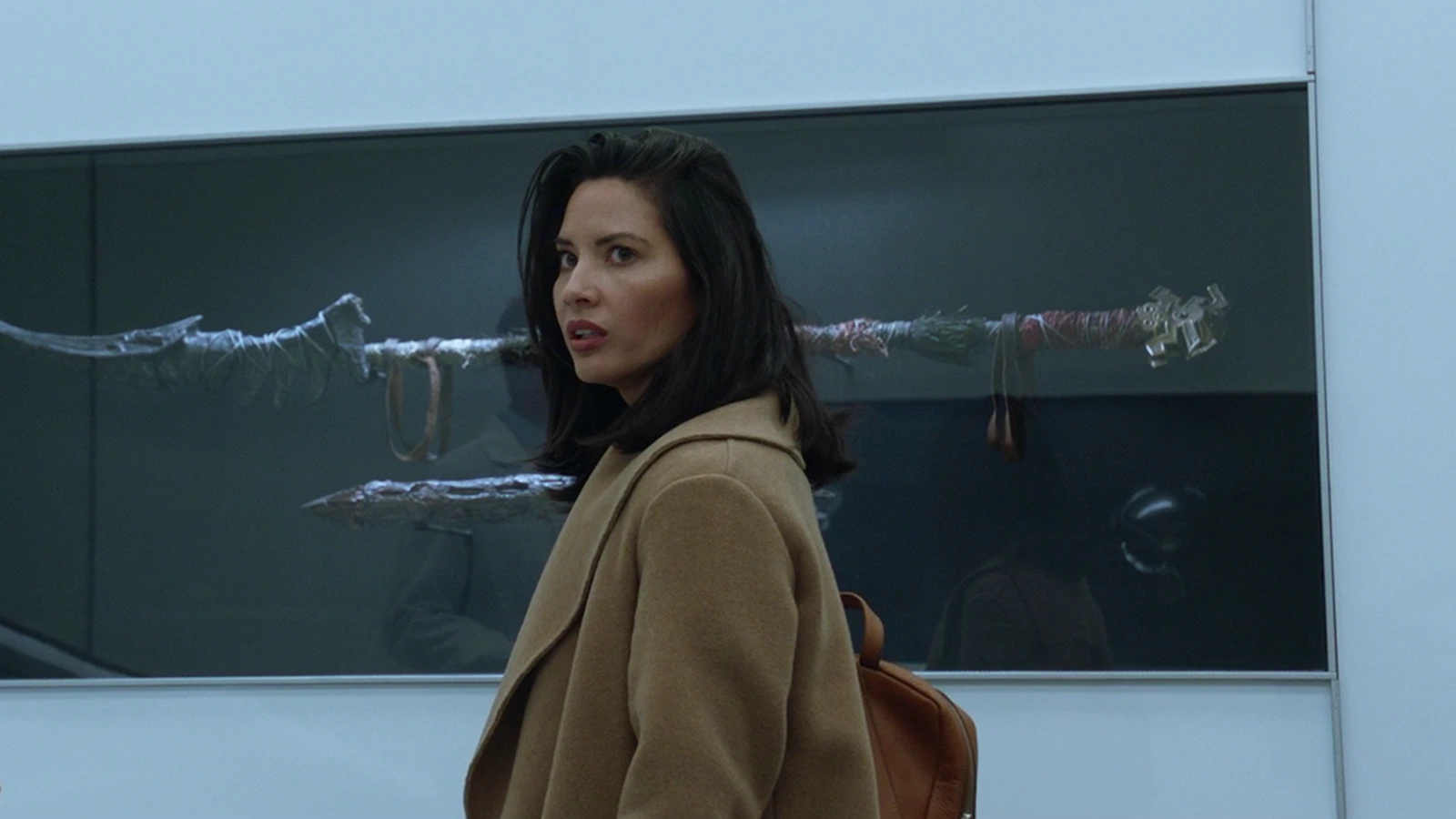 Casey Brackett in a camel coat looking back in the Stargazer lab, with Predator weapons in a display case behind her