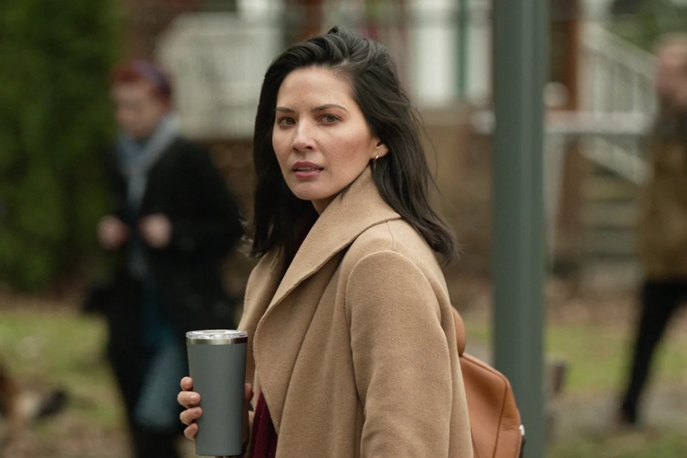 Casey Brackett in a camel coat holding a thermos on a university campus, with students walking behind her