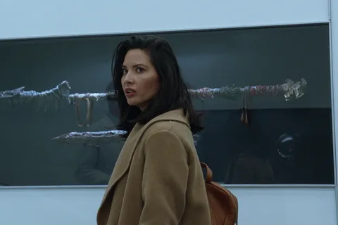 Casey Brackett in a camel coat looking back in the Stargazer lab, with Predator weapons in a display case behind her