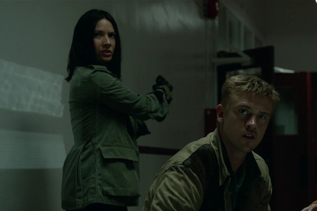 Casey Brackett aiming a pistol in a dark room while Quinn McKenna crouches beside her