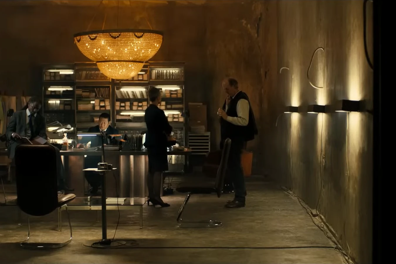 A dim concrete chamber with a long lit wall, shelves and a huge glowing chandelier above a desk in 2036: Nexus Dawn