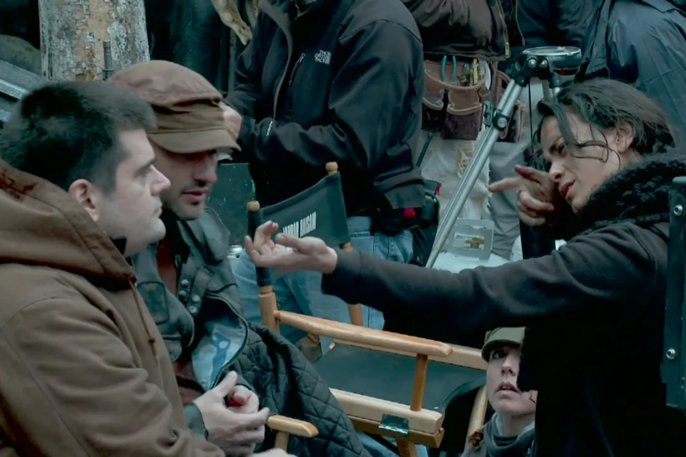 Alice Braga in a dark coat gesturing with both hands as she talks with crew members beside a director's chair on the Predators set