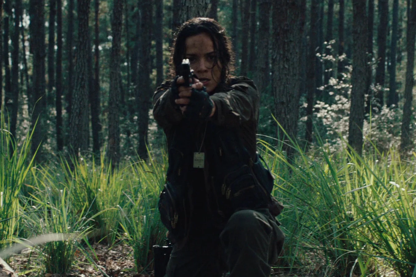 Isabelle kneeling in tall grass in a pine forest, aiming her pistol straight ahead
