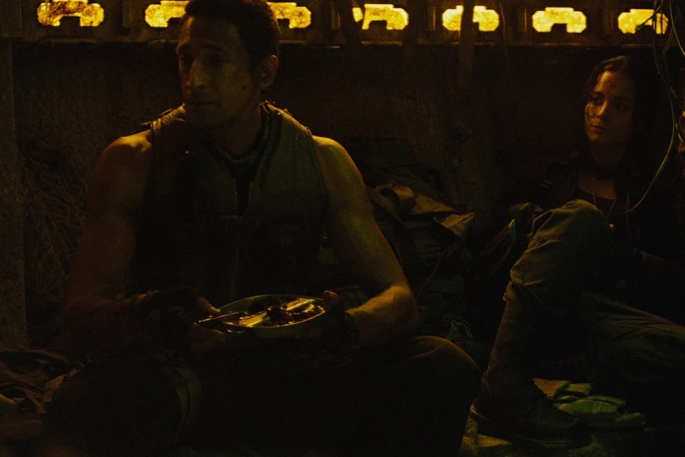 Royce sitting with a bowl of food in Noland's dimly lit hideout, with Isabelle seated behind him