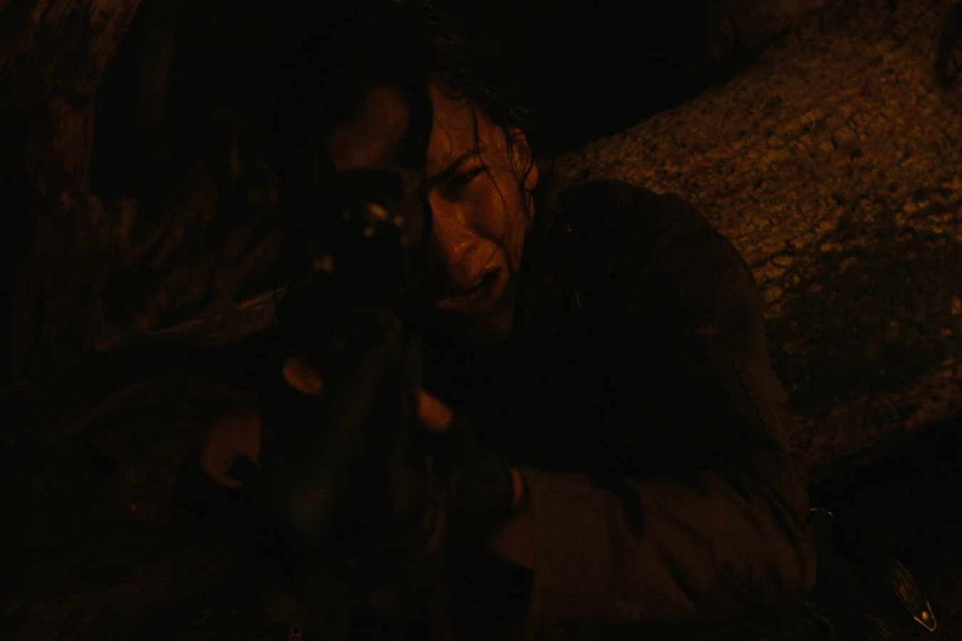 Isabelle aiming her rifle from the ground in near darkness, lit by dim amber light