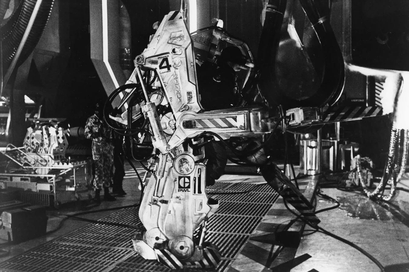 Black-and-white production photograph of the Aliens Power Loader prop on the Pinewood hangar set, marked POWER 4 LOADER and PWL