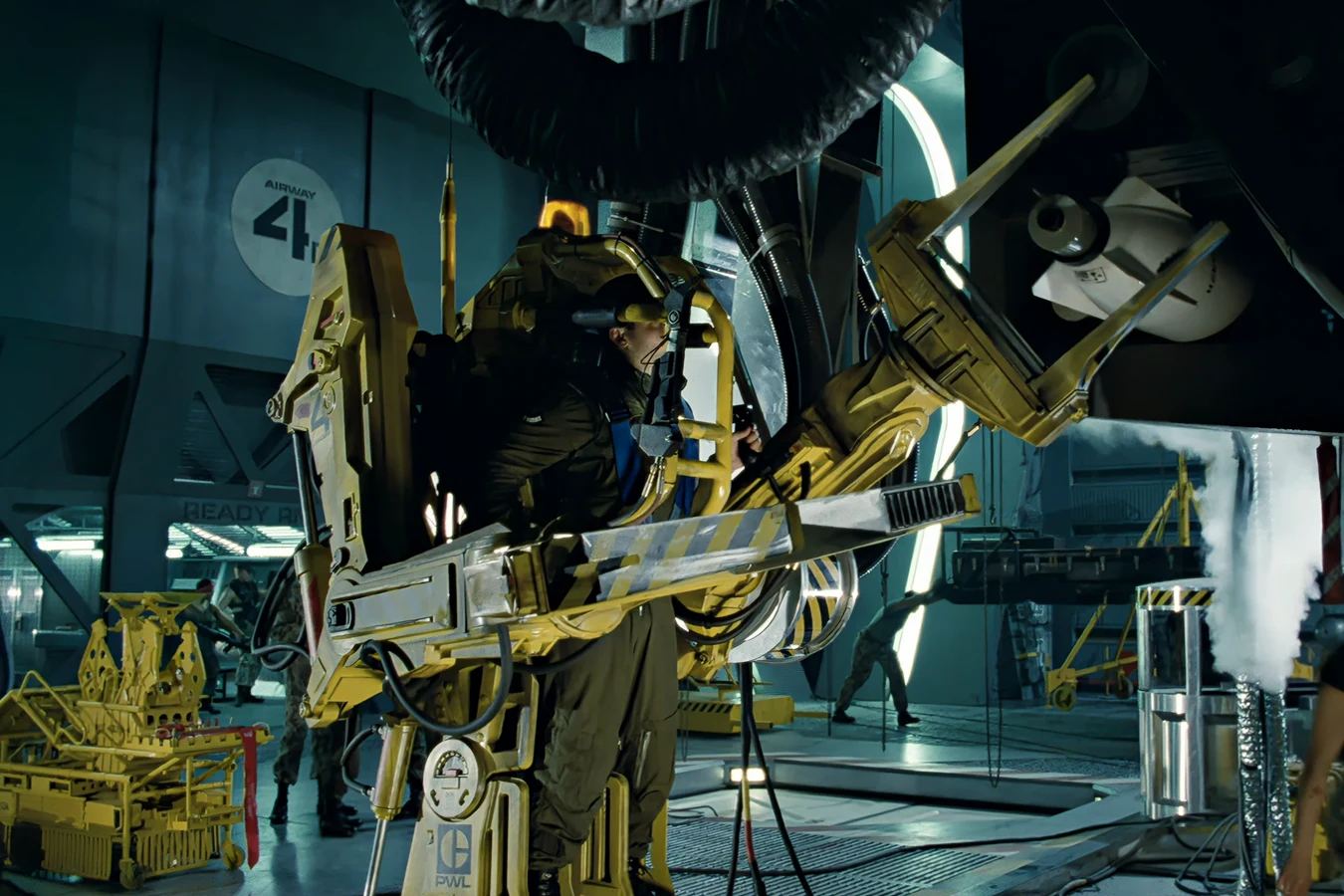 Ellen Ripley in a P-5000 Power Loader lifting white ordnance in the Sulaco hangar near the Ready Room