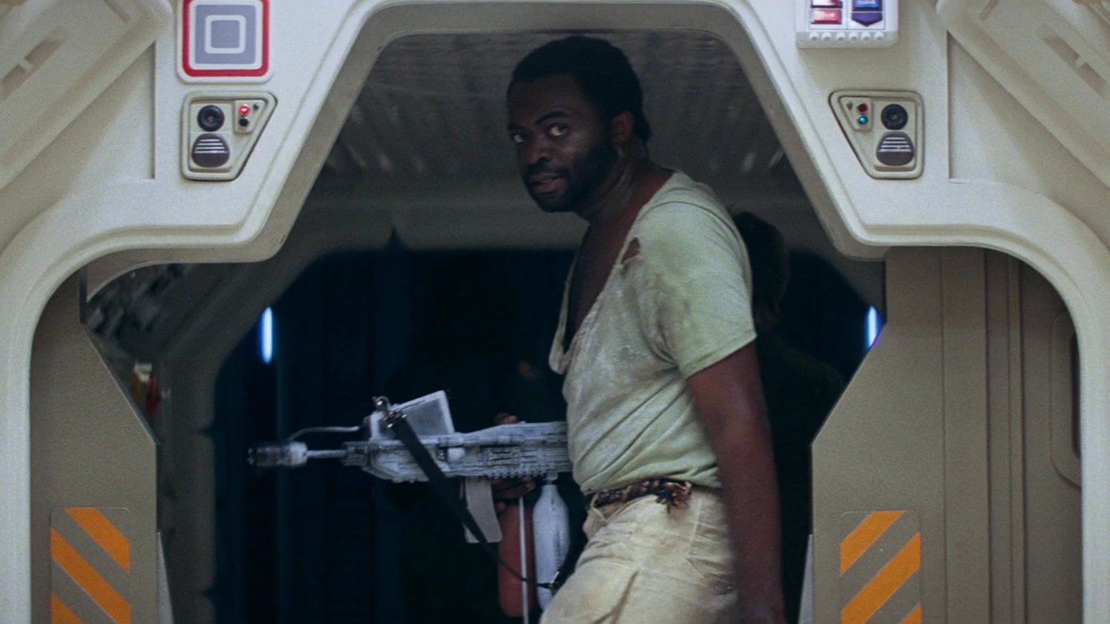 Parker in a torn undershirt carries a weapon through a Nostromo hatch in Alien