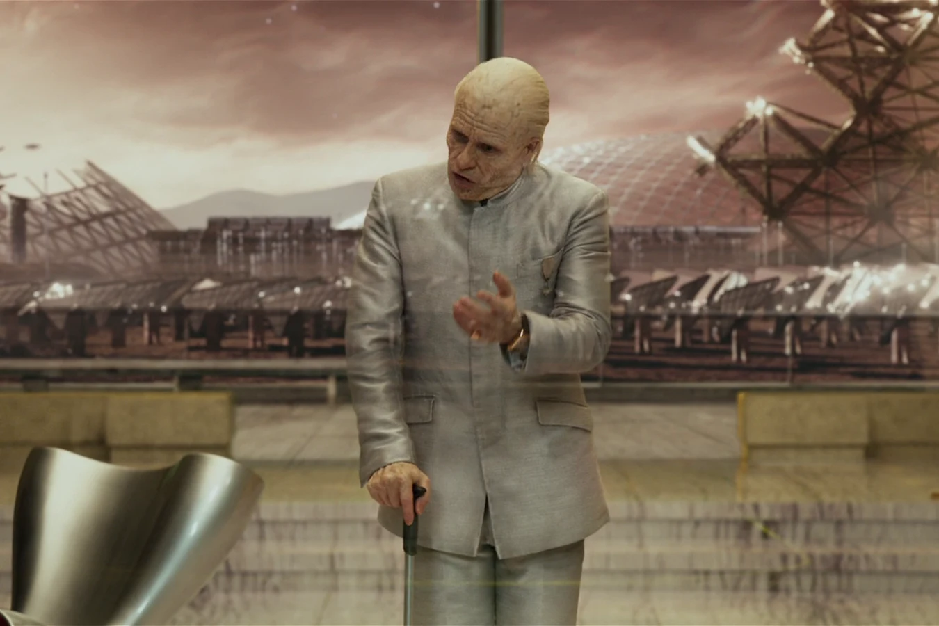 Peter Weyland with a cane in the holographic briefing, domes and towers behind the glass