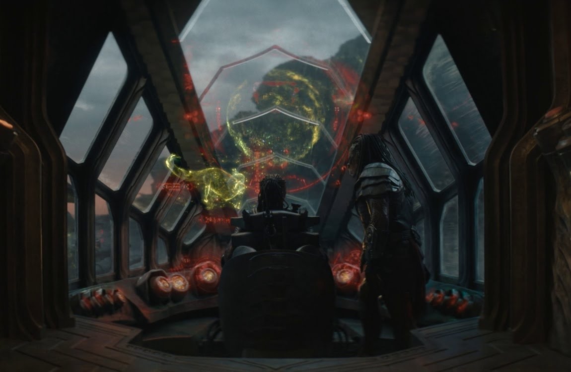 Dek and Kwei viewing a yellow planet hologram with a dinosaur-like creature projection inside Kwei's ship in Predator: Badlands