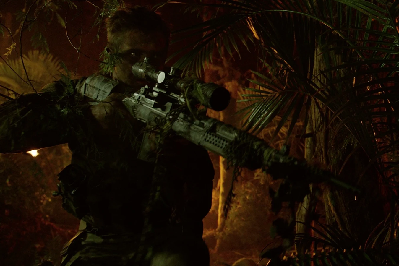 Quinn McKenna in ghillie camouflage, aiming a scoped rifle in a burning jungle