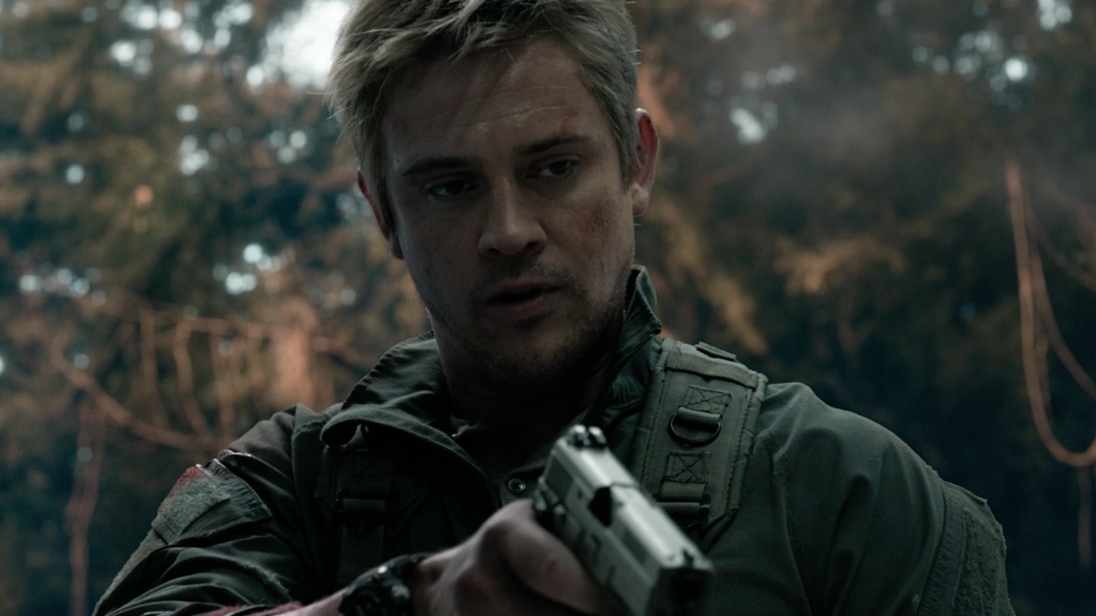 Quinn McKenna in a green field jacket, aiming a pistol in the woods with dirt and blood on his face