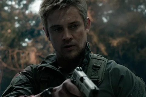 Quinn McKenna in a green field jacket, aiming a pistol in the woods with dirt and blood on his face