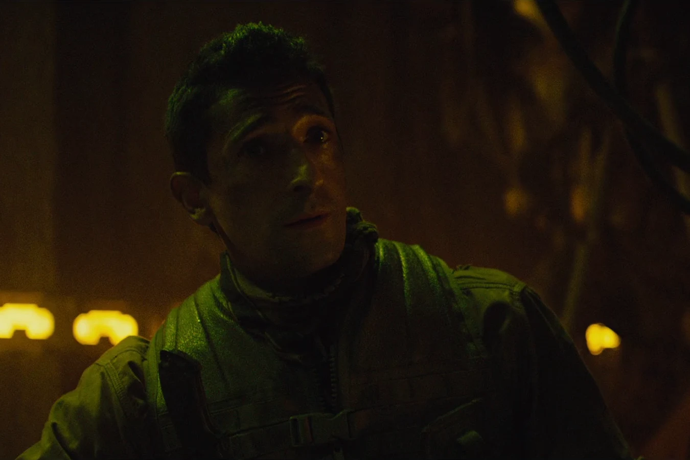 Royce in dim green light with round yellow lamps glowing behind him