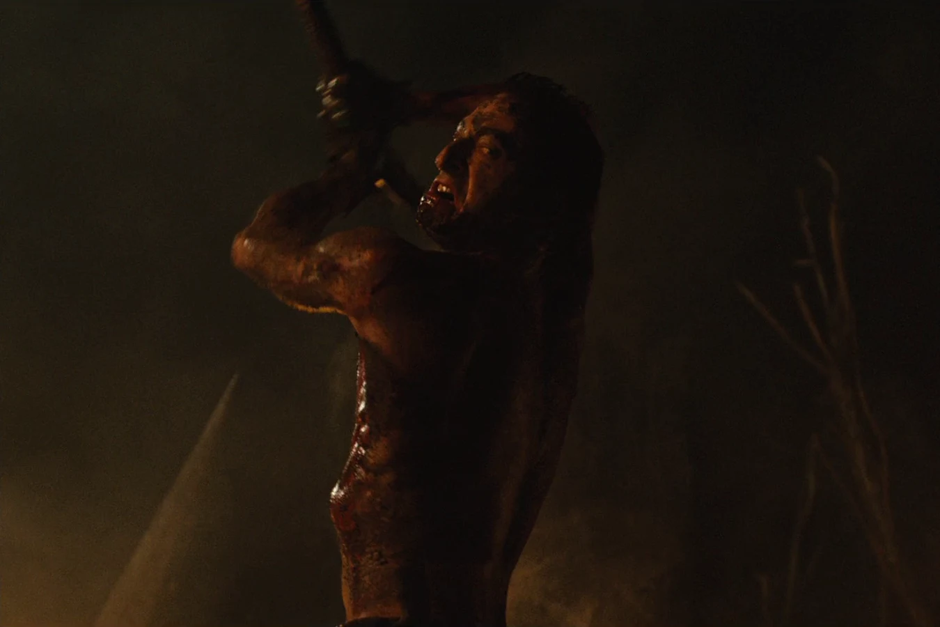 Shirtless and smeared with mud and blood, Royce silhouetted against firelight and smoke at night as he raises a weapon over his shoulder