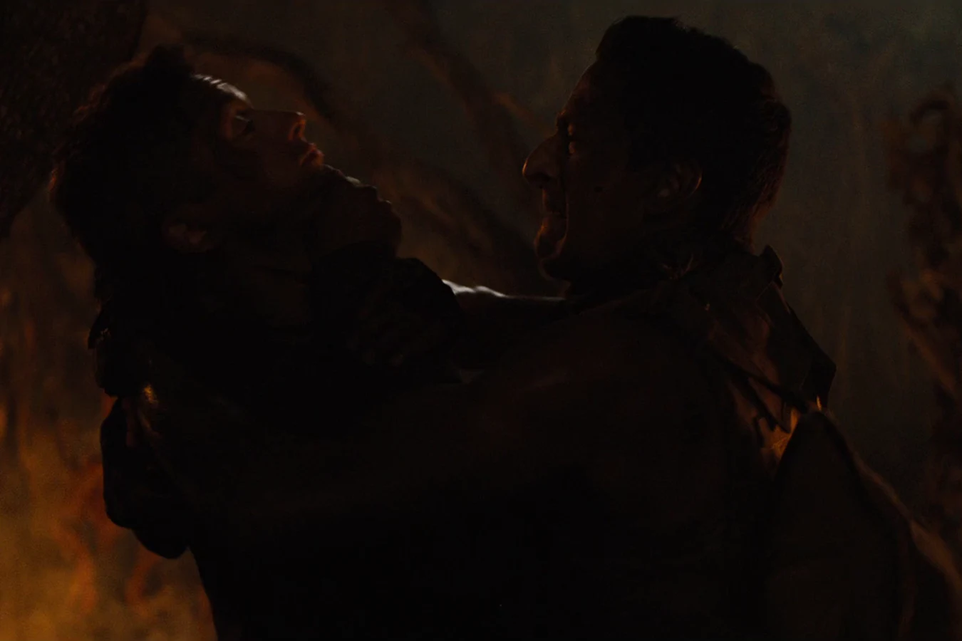 Royce gripping Edwin by the jaw and forcing his head back in the dark, with firelight glowing behind them