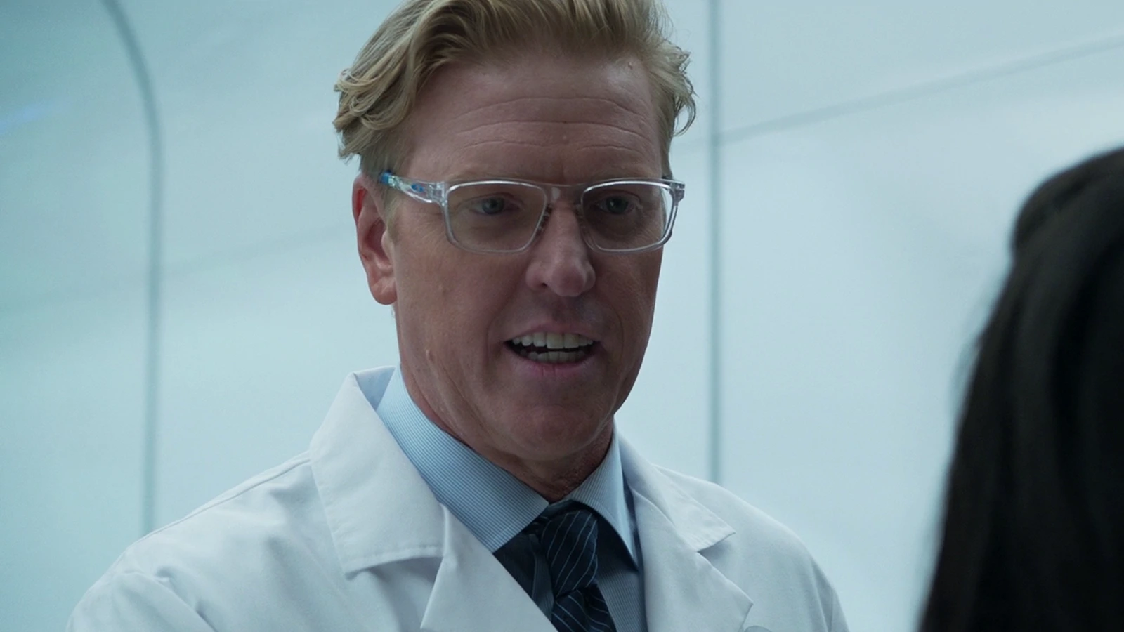 Sean Keyes in a white lab coat, clear glasses and a striped tie, talking to Casey Brackett in a white Stargazer room