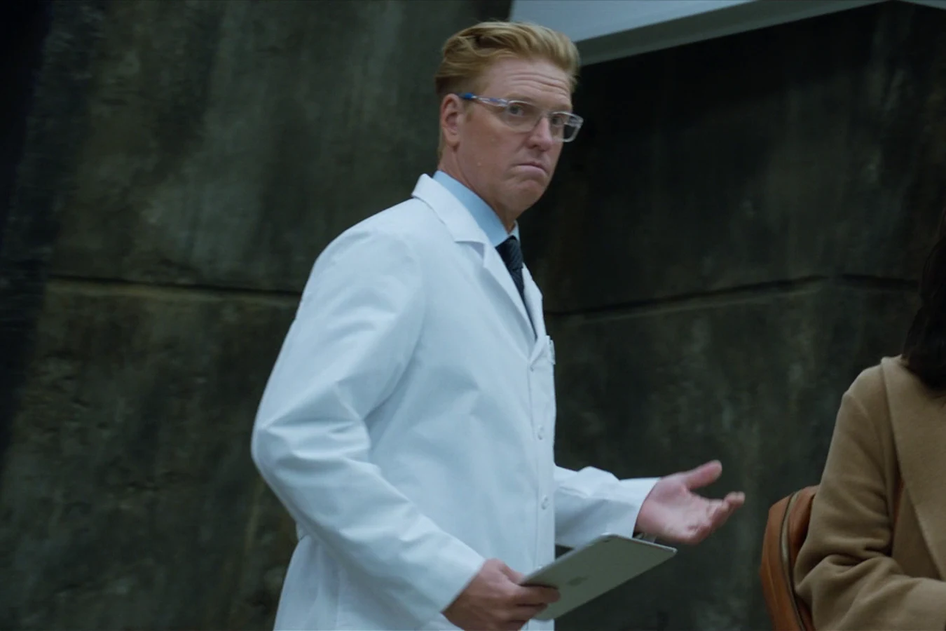 Sean Keyes in a lab coat holding a tablet and gesturing with his other hand, standing beside Casey Brackett in a tan coat and red scarf