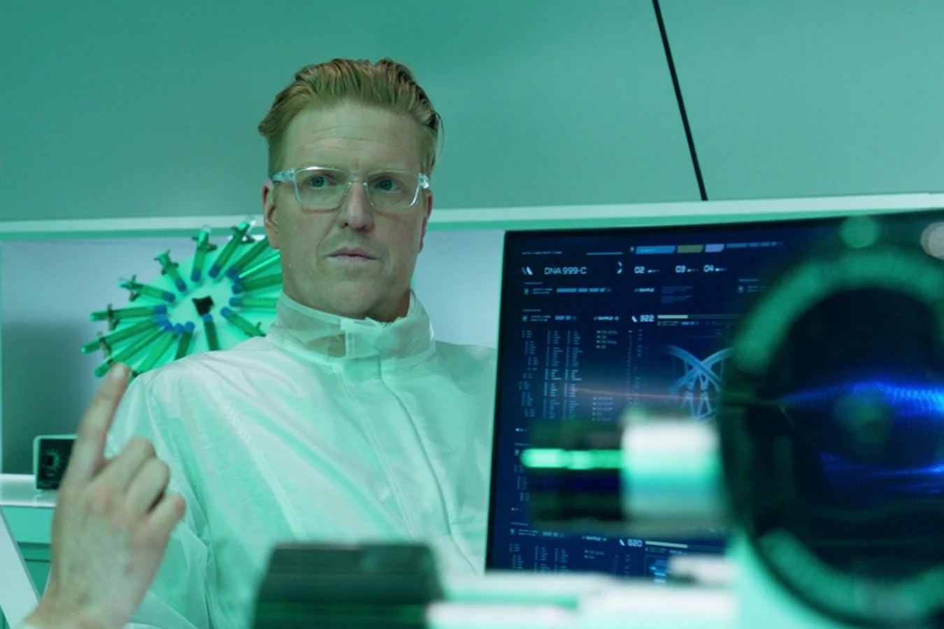 Sean Keyes in a clear protective suit at a Stargazer lab workstation beside a monitor showing a DNA readout, with Casey Brackett's raised hand in the foreground