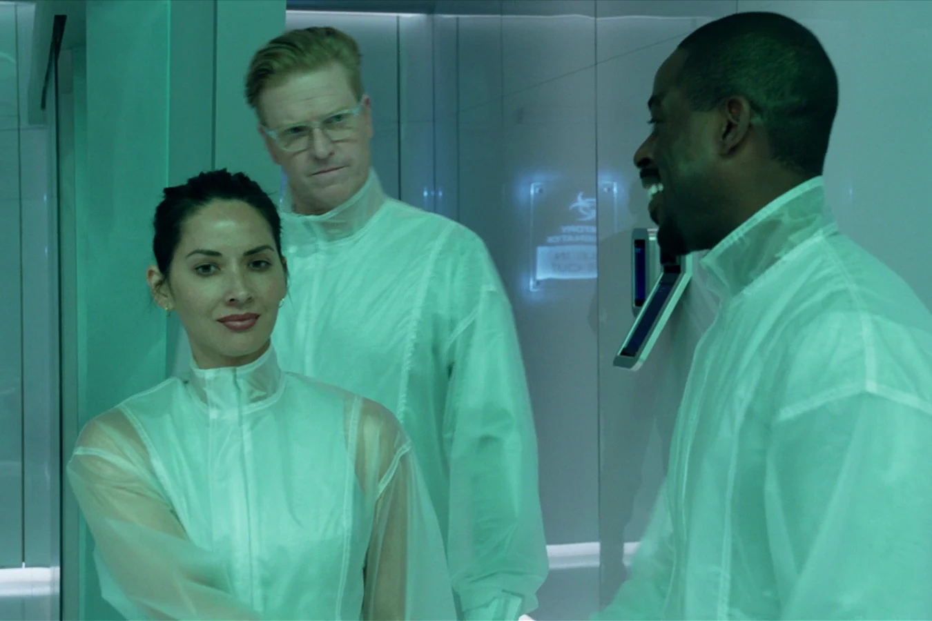 Sean Keyes in a clear protective suit standing behind Casey Brackett, with Will Traeger smiling beside them in a teal Stargazer room