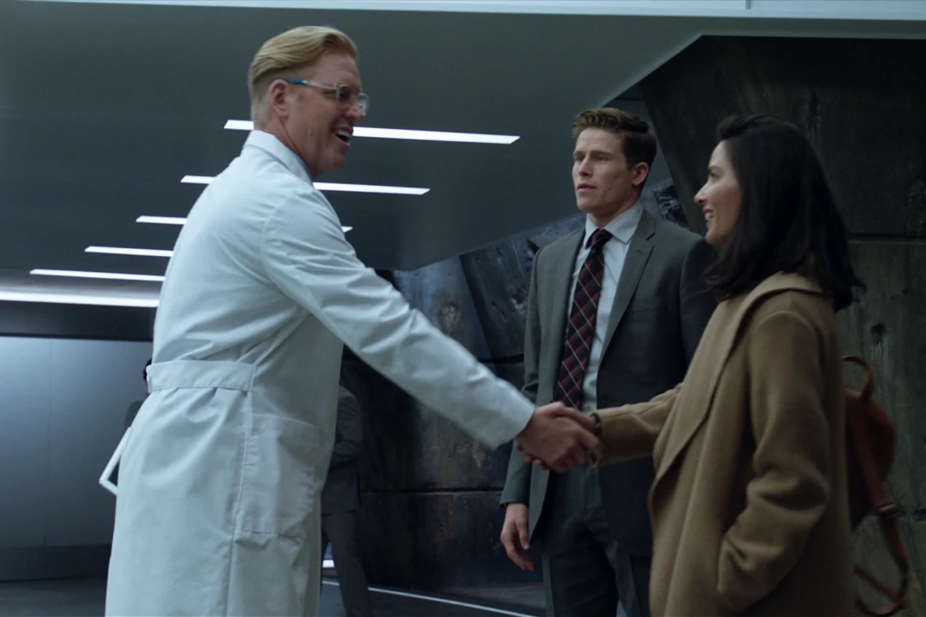 Sean Keyes in a lab coat and glasses smiling as he shakes hands with Casey Brackett in a concrete-walled Stargazer corridor, with a man in a suit behind them