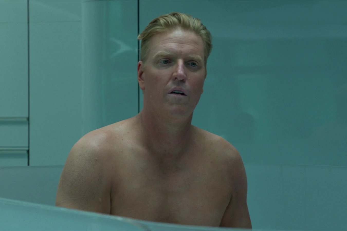 Sean Keyes shirtless behind a low partition in a teal, glass-walled room
