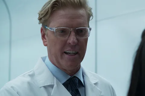 Sean Keyes in a white lab coat, clear glasses and a striped tie, talking to Casey Brackett in a white Stargazer room