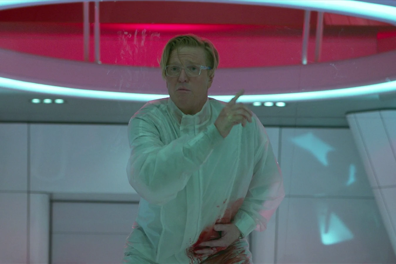 Sean Keyes in a protective suit with blood at his waist, pointing upward under a red-lit ring in the ceiling of the lab