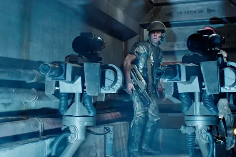Hudson standing between two UA 571-C Automated Sentry Guns in a Hadley's Hope corridor